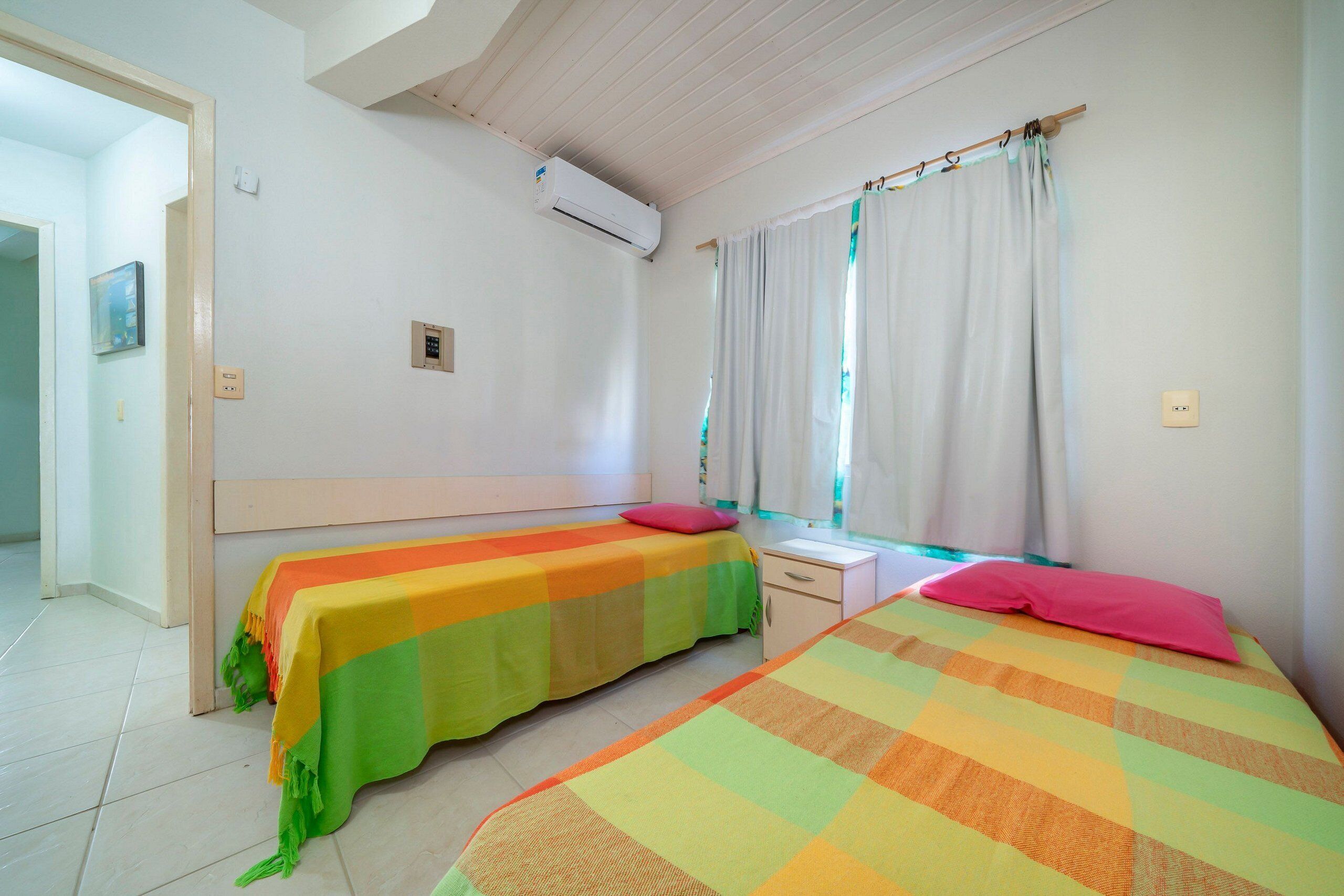 Ein Zimmer mit zwei Betten, jeweils mit bunter Bettwäsche, einem kleinen Nachttisch und einem Fenster mit Vorhängen.