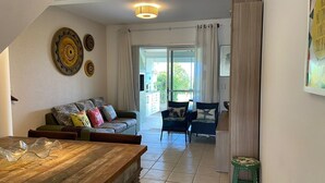 A living room with a wooden dining table, a sofa with patterned cushions, and two armchairs. A large window with curtains is visible, leading to an outdoor area. - Residencial Guilhermina (Bombinhas)