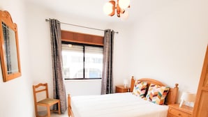 A bedroom with a bed, a chair, a dresser, a nightstand, a lamp, a window with curtains, and a ceiling fan. - T1 Largo Frente - Familiar (Loulé)