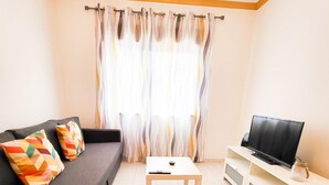 A living room with a sofa, a coffee table, a TV on a stand, and a window with curtains. - T1 Largo Frente - Familiar (Loulé)