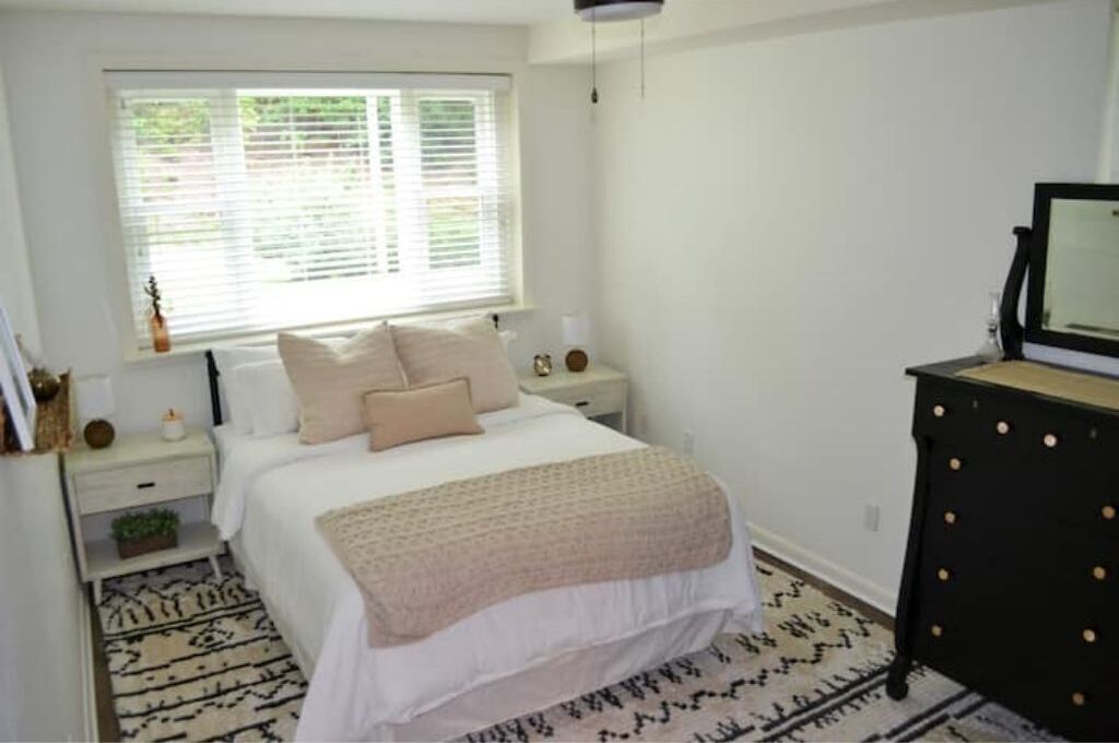 Un dormitorio con cama, mesitas de noche, cómoda, ventana con persianas y una alfombra estampada.