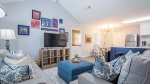 Een moderne woonkamer met een flatscreen televisie, een bank, een poef en een eethoek, met een keuken op de achtergrond.