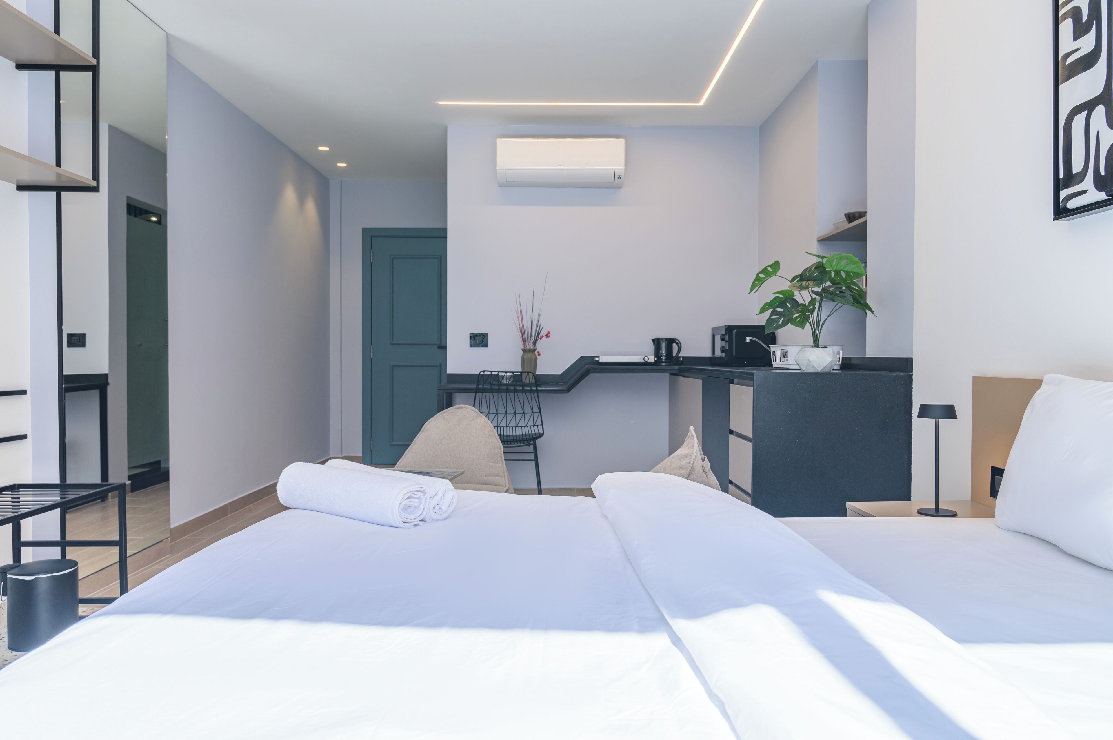 A modern hotel room with two beds, a kitchenette, a desk, and a potted plant.