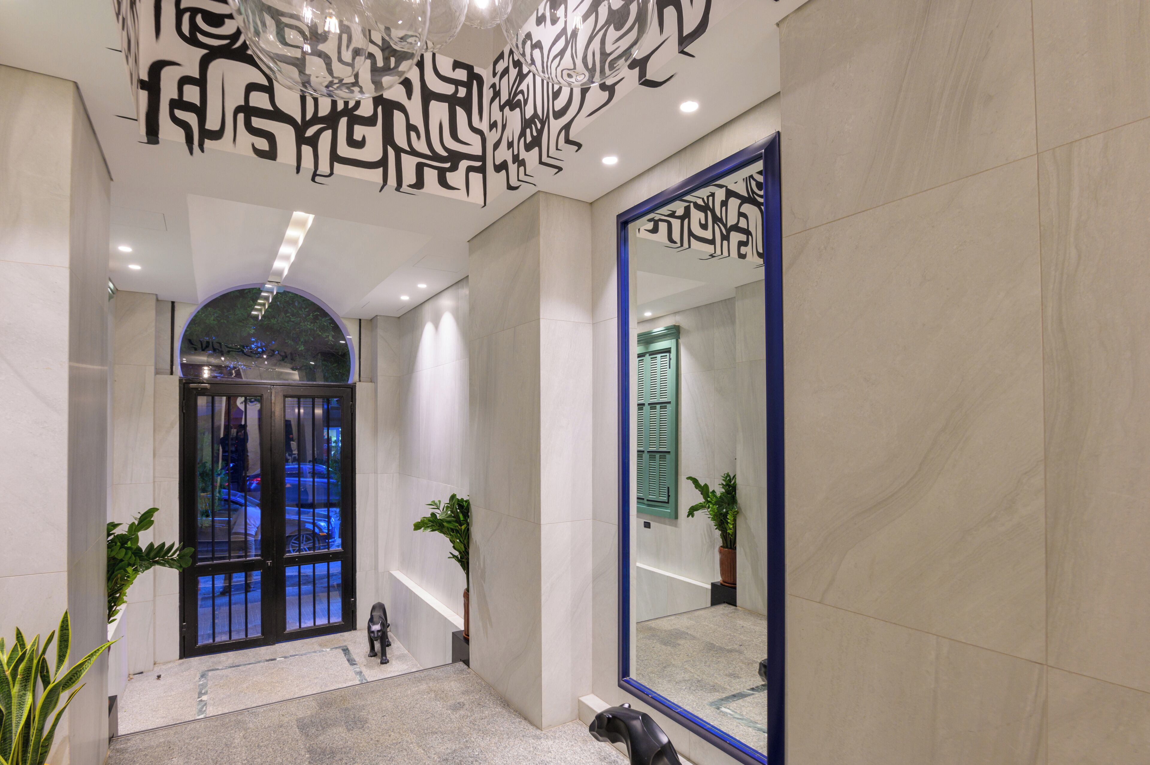 A modern hotel lobby with a large mirror, a glass door entrance, and potted plants.