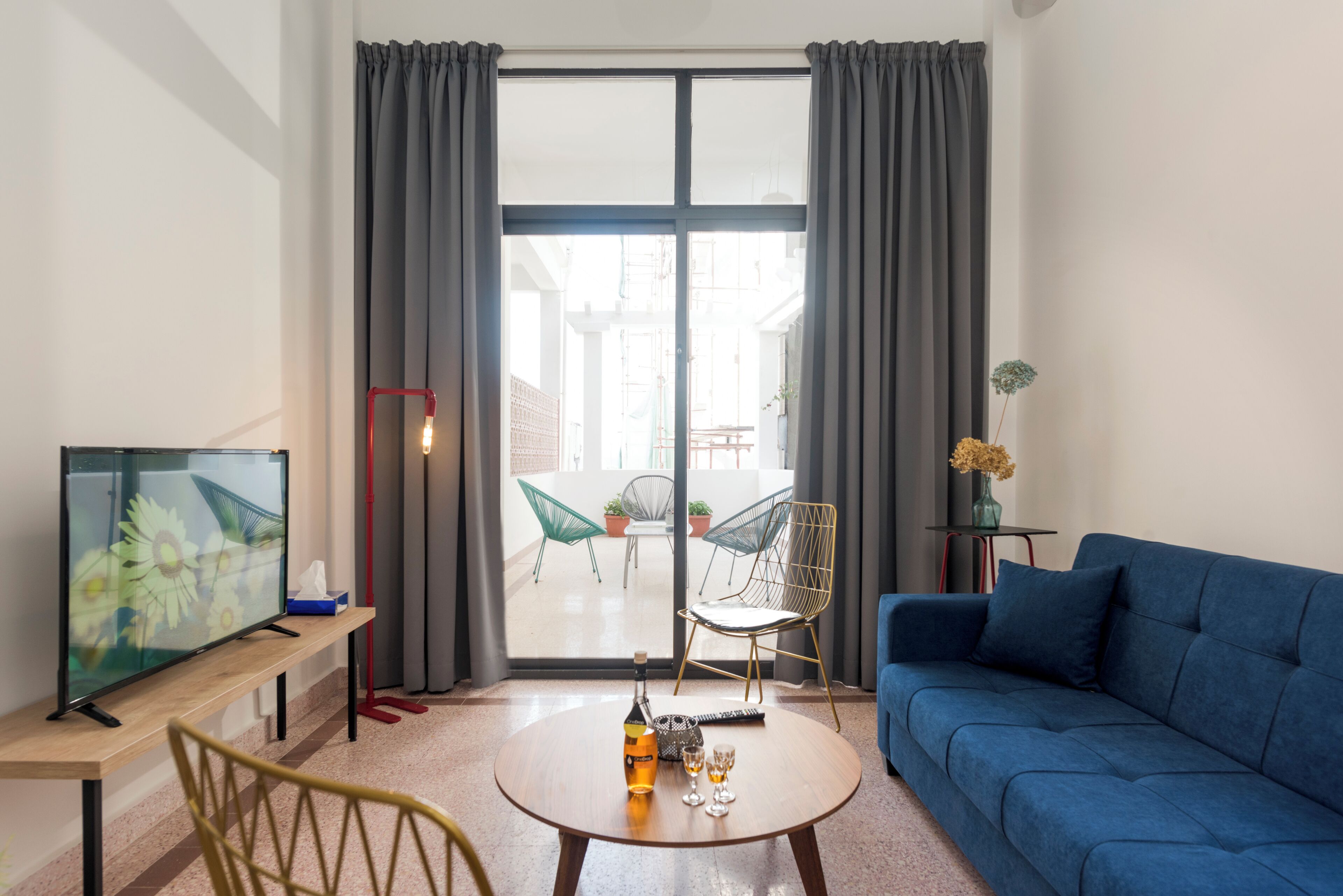 A modern living room with a blue sofa, a wooden coffee table with a bottle and glasses, a TV on a wooden stand, and a view through large glass doors to an outdoor patio with chairs and plants.
