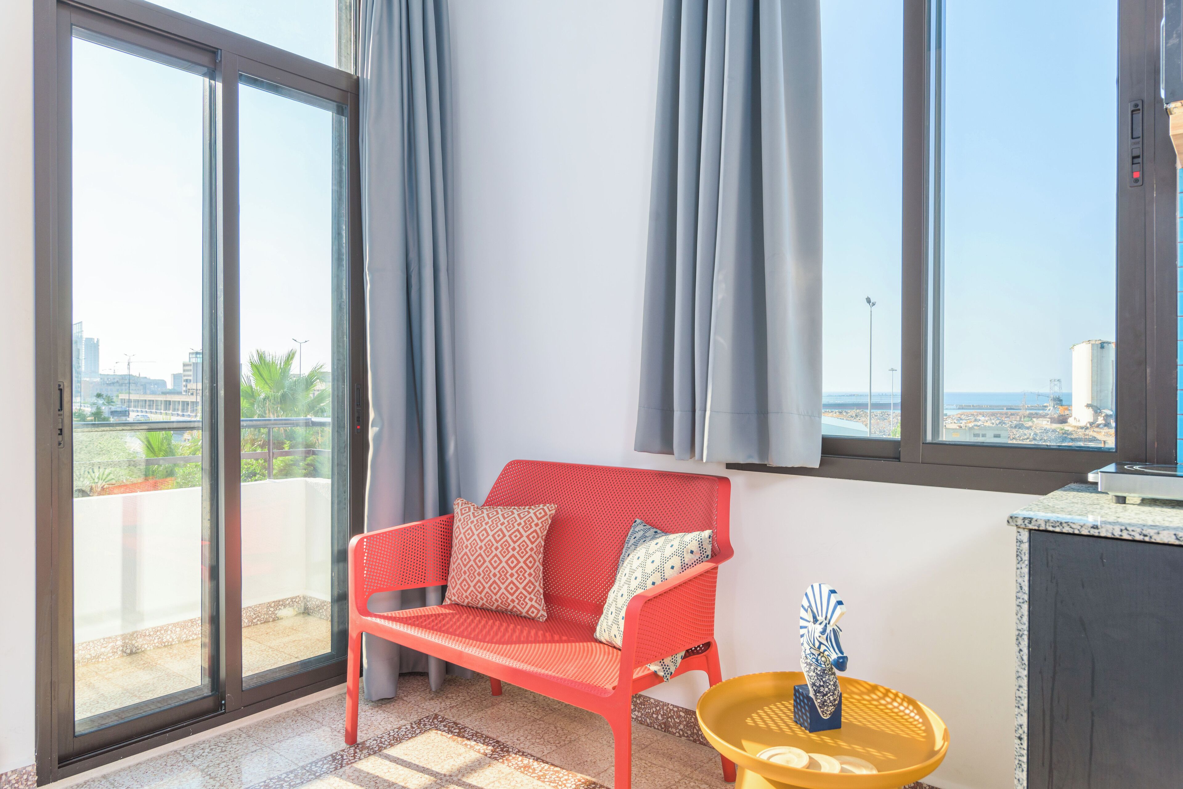 Una habitación con un sofá rojo, una pequeña mesa amarilla y vistas de la ciudad a través de grandes ventanales.