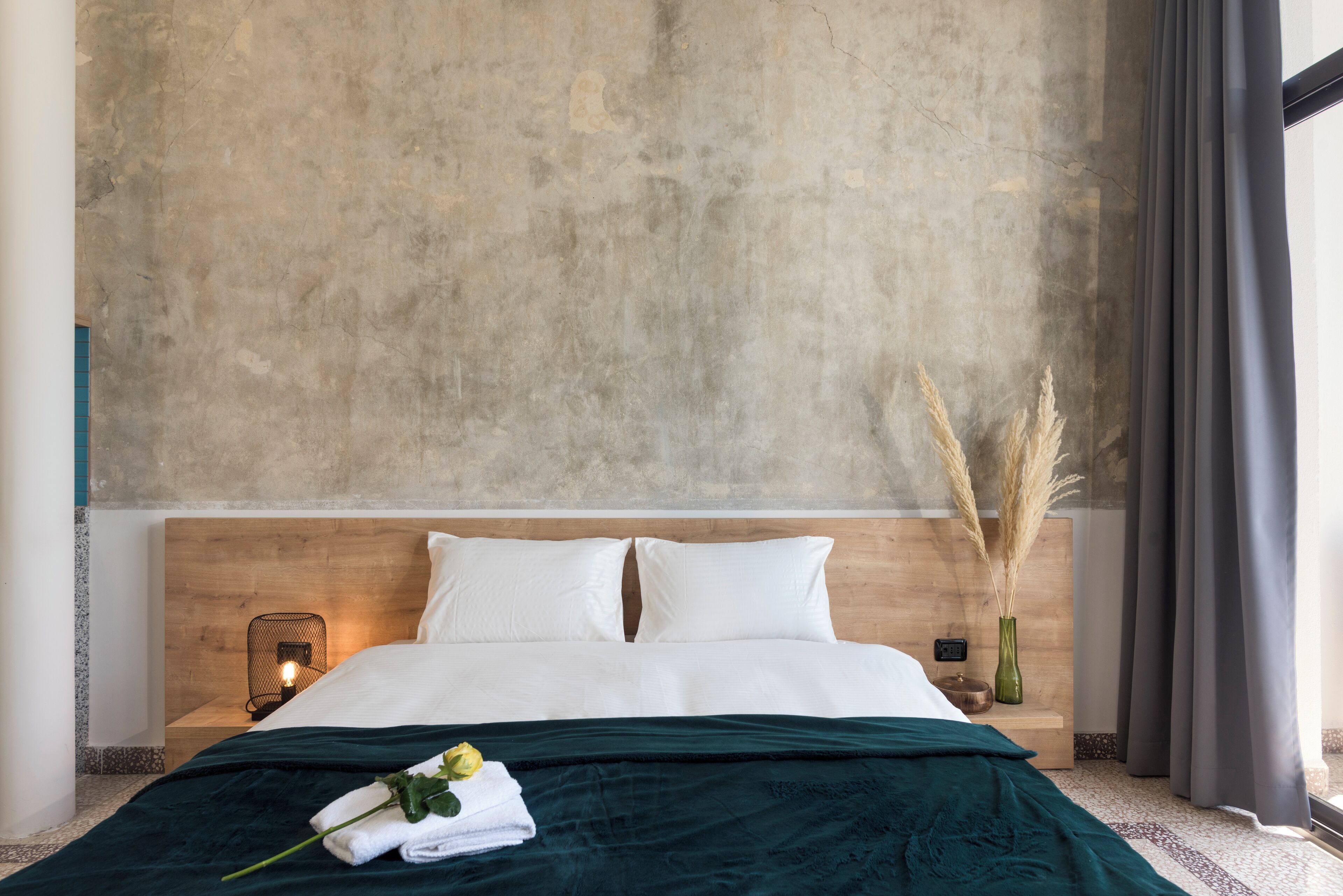A modern hotel room with a large bed, a wooden headboard, a nightstand with a lamp, and a vase with dried plants.