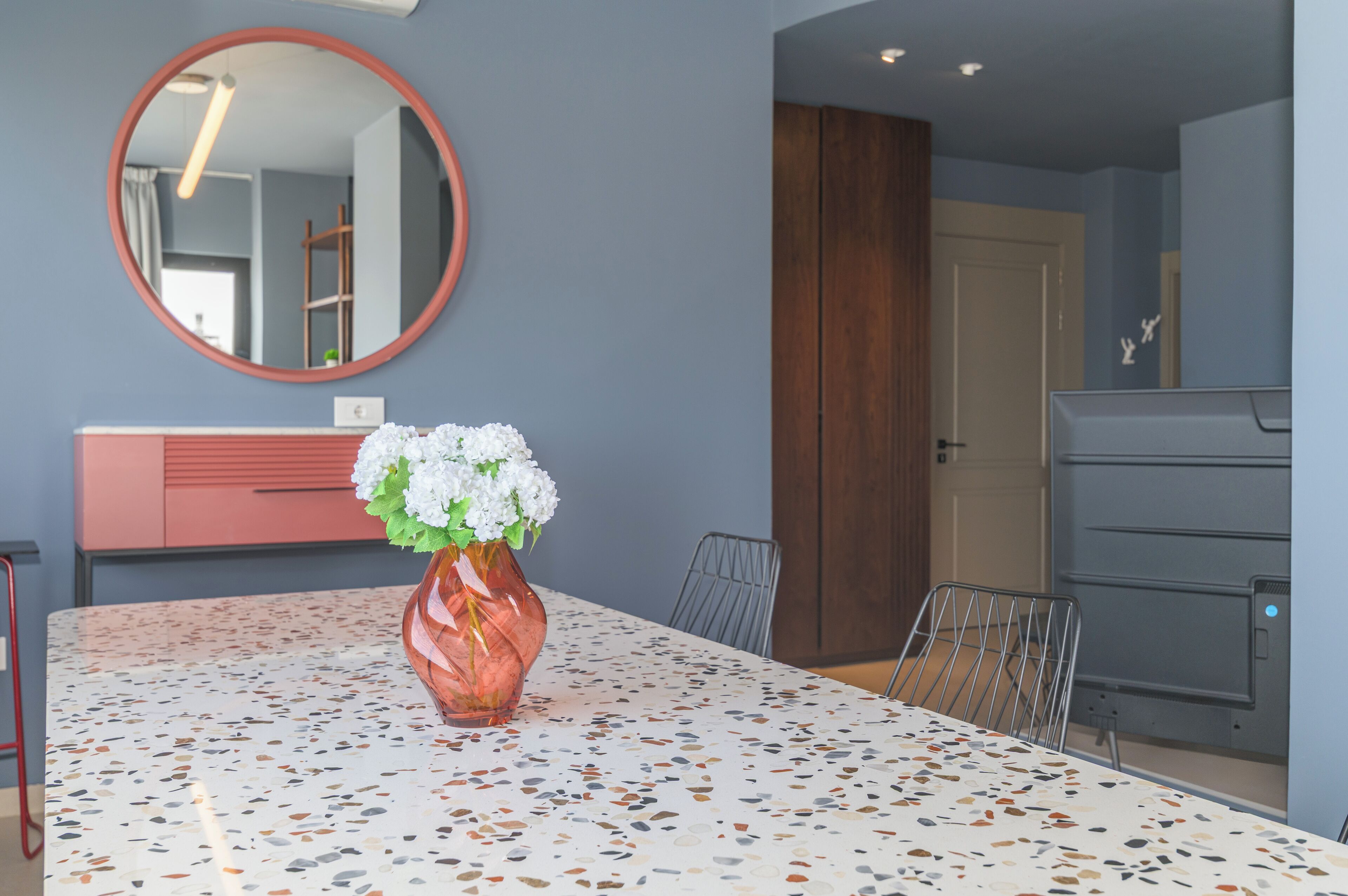 A modern dining area with a terrazzo table, a vase of white flowers, a round mirror, and a wooden cabinet.