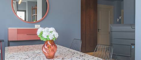 Ein stilvoller Essbereich mit einem Terrazzo-Tisch, einer Vase mit weißen Blumen, einem runden Spiegel und einem Holzschrank.