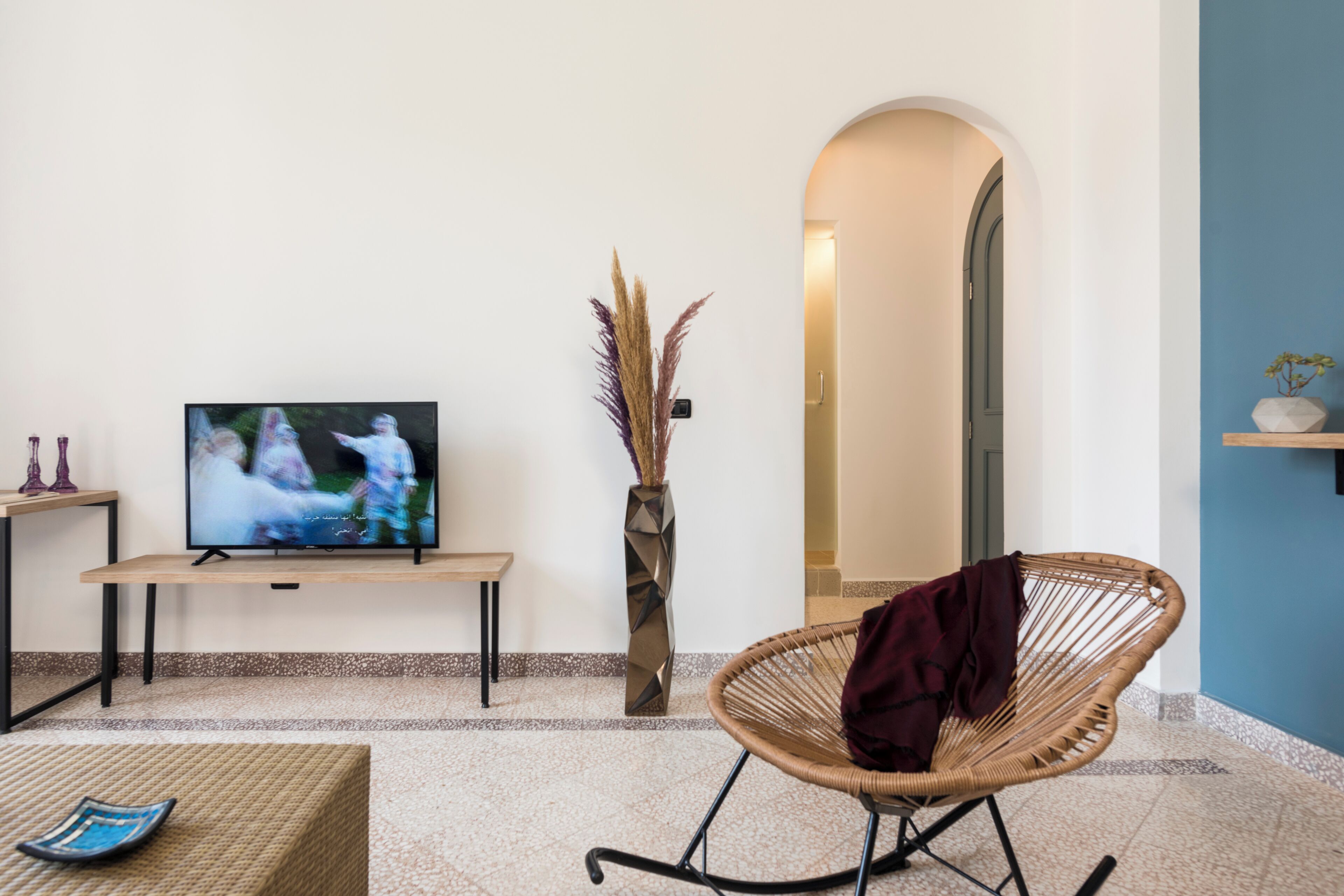 A living room with a television, a wicker chair, a vase with dried plants, and a small table with a blue object.
