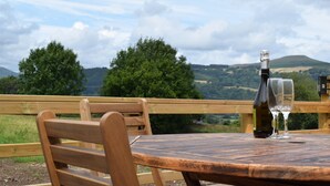 Uma mesa de madeira com uma garrafa de vinho e um copo, com vista para uma paisagem cênica com árvores e colinas.