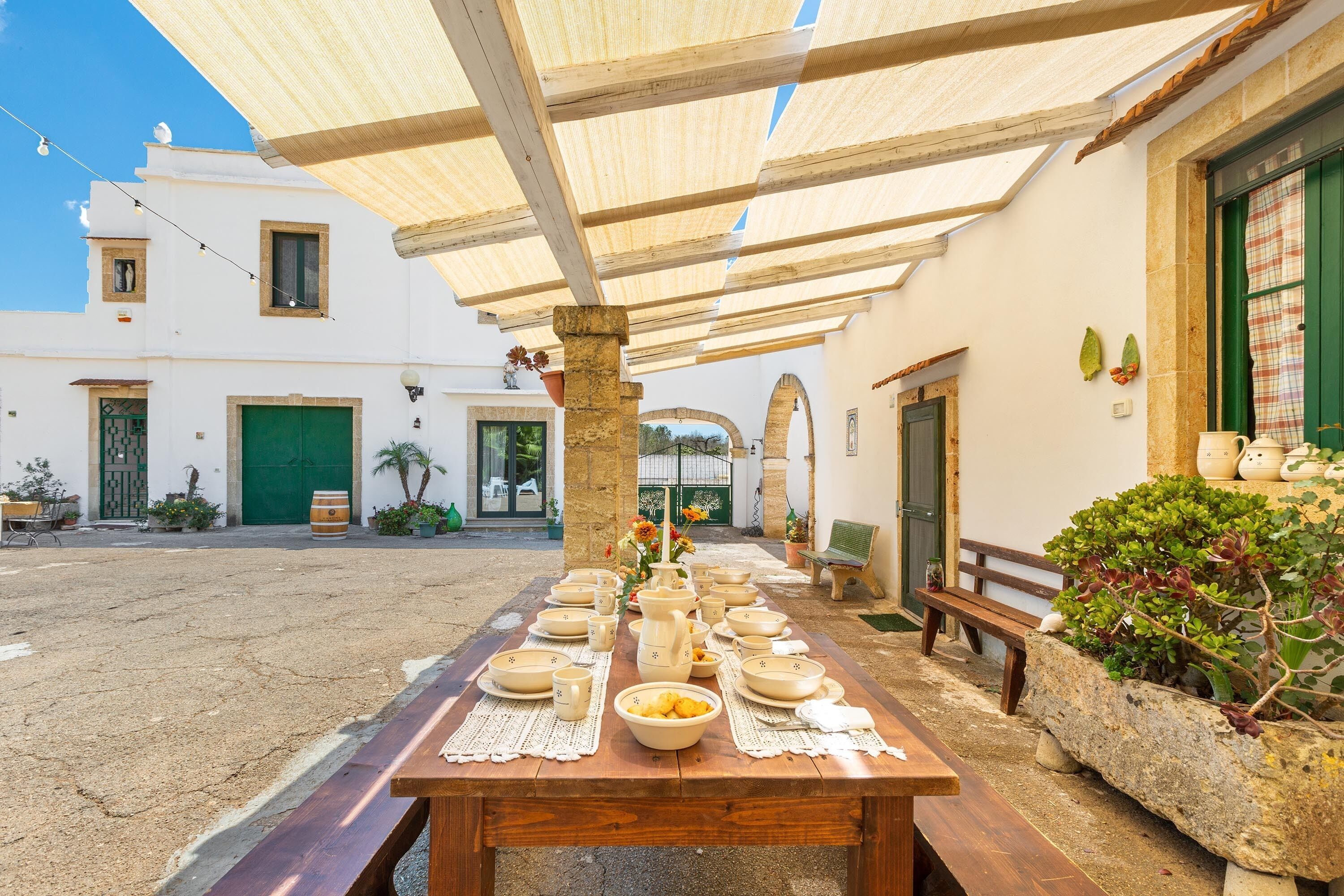 Un área de comedor al aire libre con una larga mesa de madera dispuesta con platos, tazones y tazas, rodeada por un porche cubierto con un pilar de piedra y un banco.