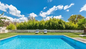 Una piscina rettangolare con acqua cristallina, circondata da un prato curato e un siepe. Due lettini con cuscini a righe sono rivolti verso la piscina.