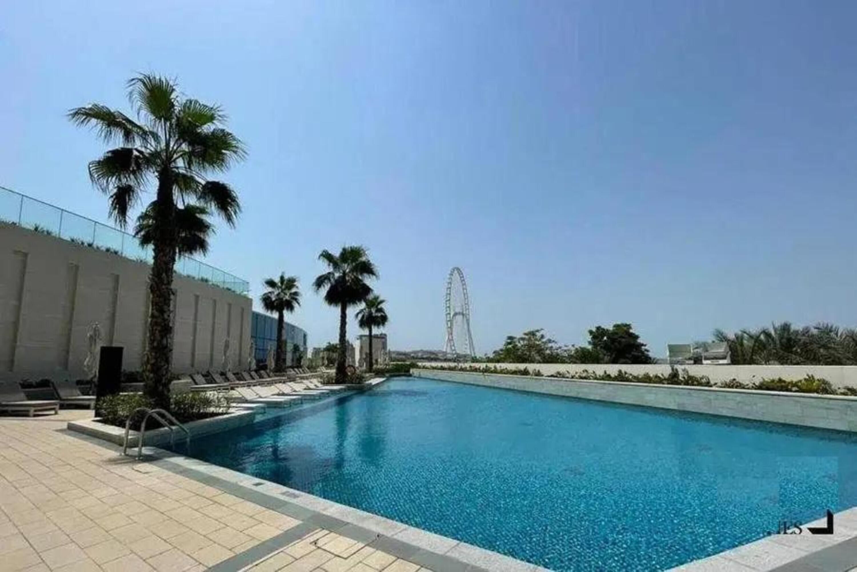 A swimming pool with lounge chairs and palm trees, a modern building, and a Ferris wheel in the background.