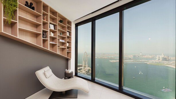 A modern room with a curved white chair, a large window with a view of a city and water, and a wooden shelving unit with various items. - Maison Privee - Lavish Apt w/ Stellar Ocean Views in Address JBR (Dubai)