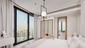 A modern hotel room with a large bed, a bedside table, a nightstand, a desk, a chair, a large window with a city view, and a door leading to a balcony. - Maison Privee - Lavish Apt w/ Stellar Ocean Views in Address JBR (Dubai)