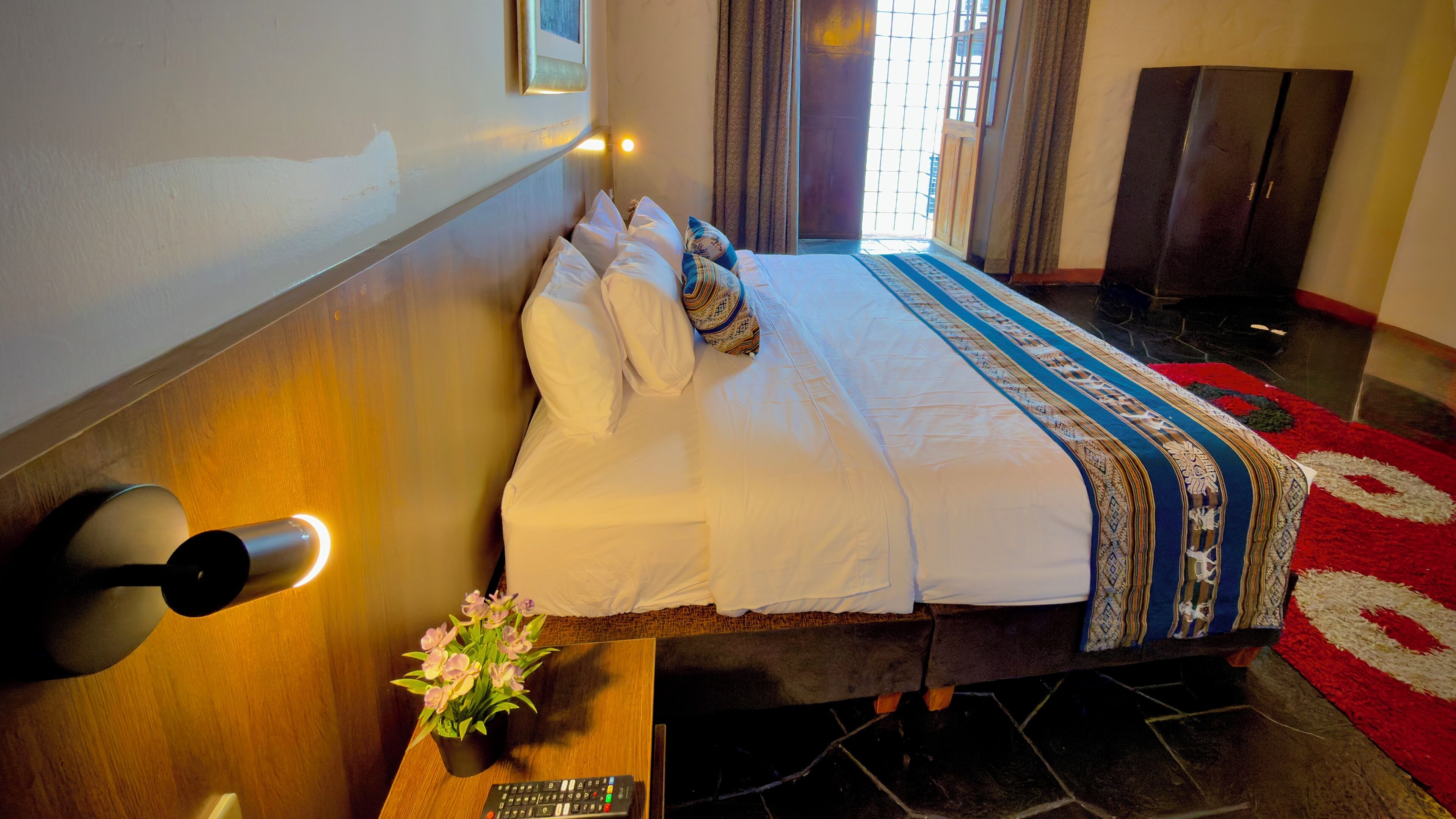 A neatly made bed with white linens and patterned pillows, a bedside table with a remote control and a potted plant, and a red carpet with a geometric design.