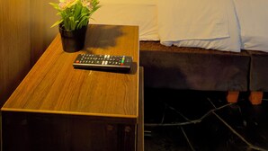 Een hotelkamer met een houten hoofdeinde, een bed met witte lakens, een nachtkastje met een afstandsbediening, en een raam met gordijnen.