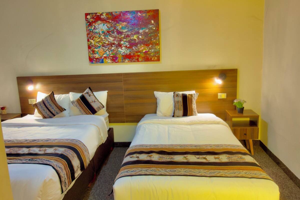 Een hotelkamer met twee bedden, een houten hoofdbord, een kleurrijk abstract schilderij en een kleine tafel met een plant.