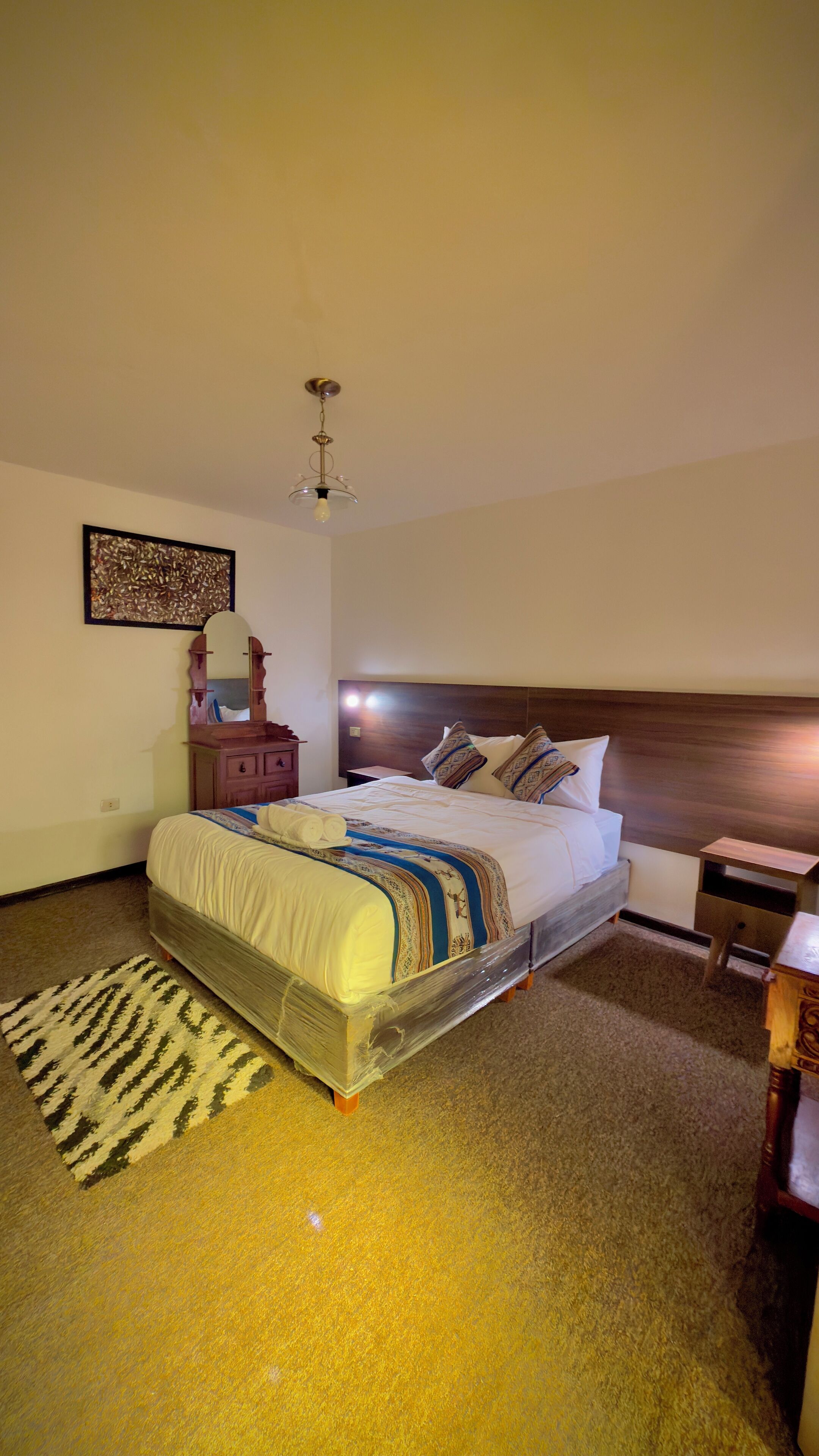 A neatly arranged hotel room with a large bed, a wooden headboard, a bedside table, a nightstand, a mirror, and a patterned rug.