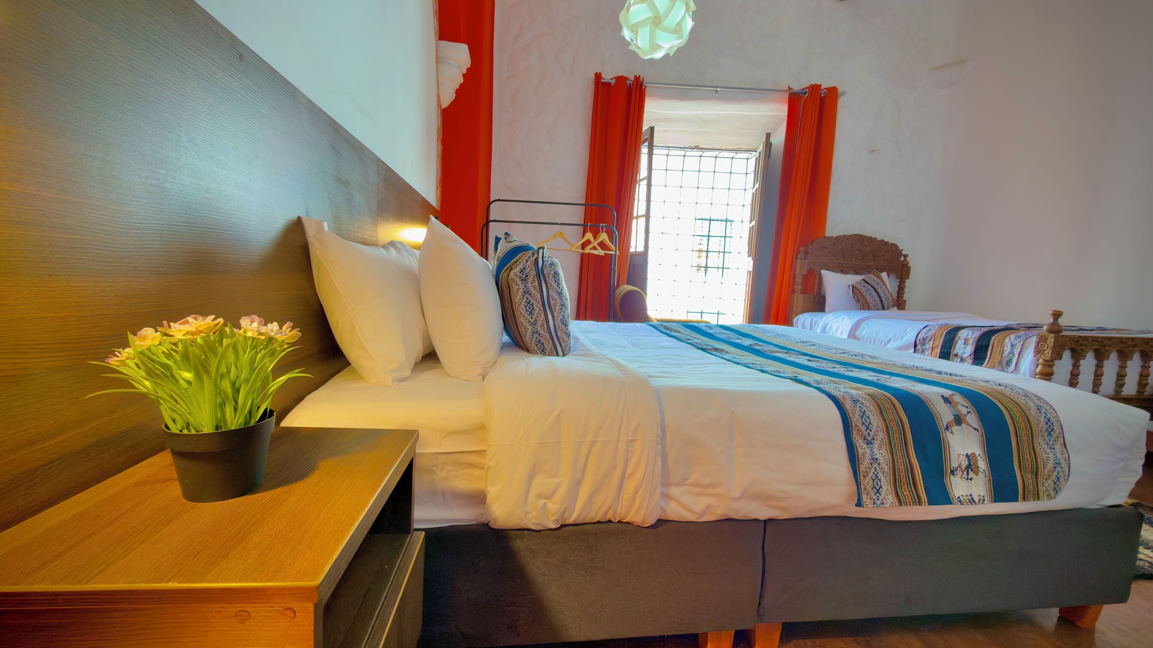 A hotel room with two beds, a wooden headboard, a bedside table with a plant, and a window with curtains.