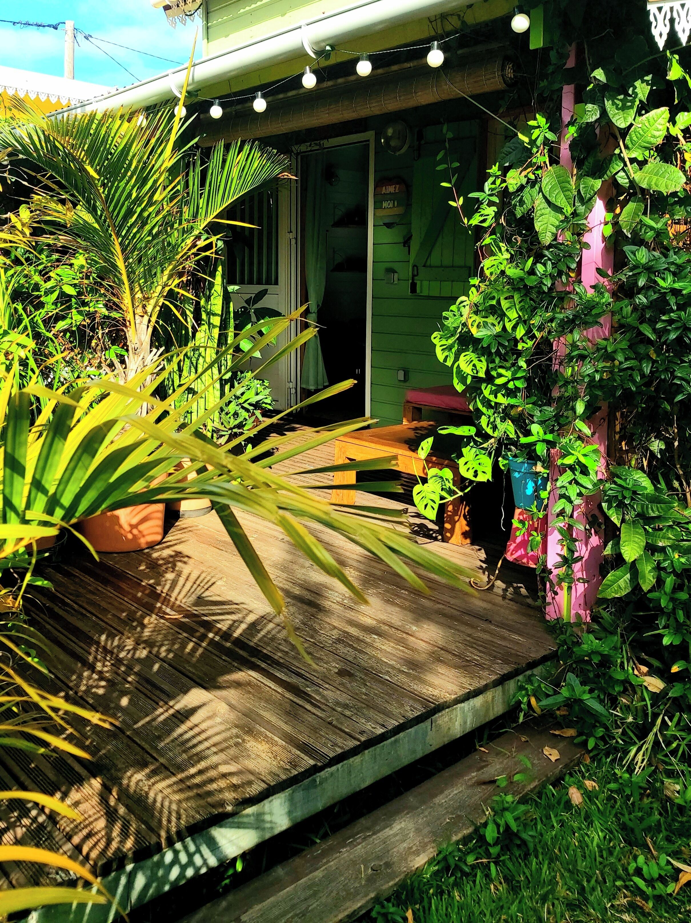 Una terraza de madera con macetas y un banco, que conduce a una casa con una puerta verde y una ventana.