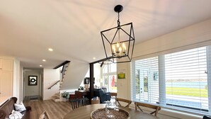 A spacious dining area with a wooden table, chairs, and a woven centerpiece. A staircase leads to an upper level with a view of a body of water. - Water View Cottage With Access To River & Minutes To Sandy North Shore Beaches (Kensington)