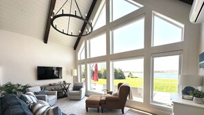 A spacious living room with a large sectional sofa, a brown armchair, and a dining area with a table and chairs. - Water View Cottage With Access To River & Minutes To Sandy North Shore Beaches (Kensington)