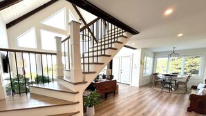 A spacious living area with a wooden staircase, a dining table with chairs, and a large window providing natural light. - Water View Cottage With Access To River & Minutes To Sandy North Shore Beaches (Kensington)