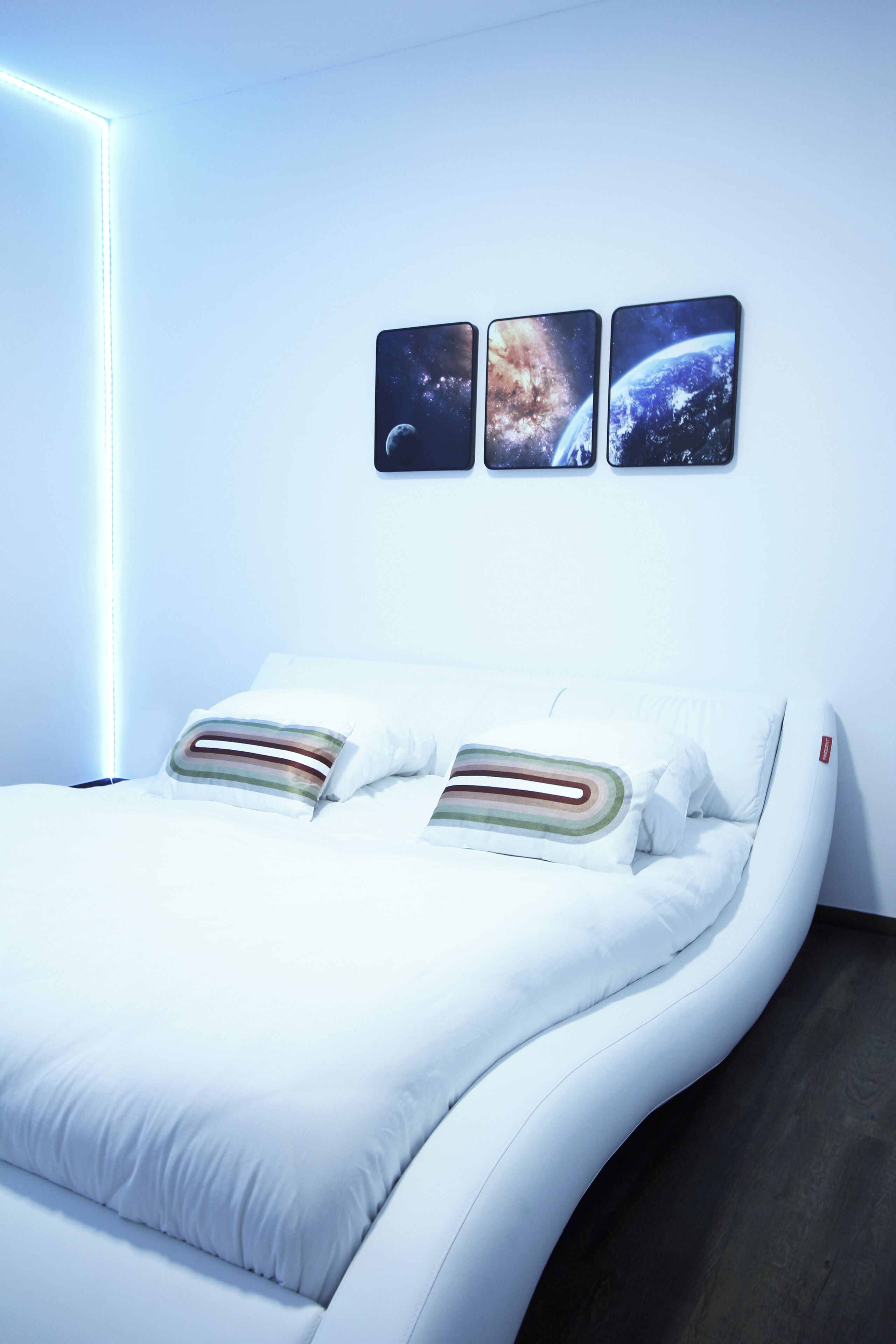 Une chambre à coucher moderne avec un lit aux lignes courbes, une literie blanche et trois tableaux encadrés sur le thème de l’espace accrochés au mur.