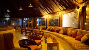 Un espacio lounge acogedor con un sofá curvo, sillas de mimbre y una mesa de madera. Amplias ventanas ofrecen vistas del exterior.