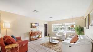 A living room with a red sofa, white armchair, and a wooden side table. There is a large window with blinds, a painting on the wall, and a small plant. - Waterfront-2BDR/2BTH -4 mins to the Beach+ Kayaks (Pompano Beach)