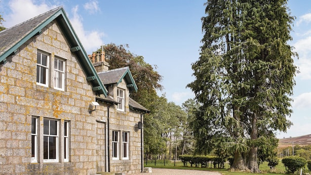 Ein Haus aus Stein mit einem grünen Dach, großen Fenstern und einer Auffahrt mit Kies.
