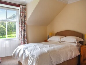 A bedroom with a bed, a window with curtains, a nightstand with a lamp, and a view of greenery outside.
