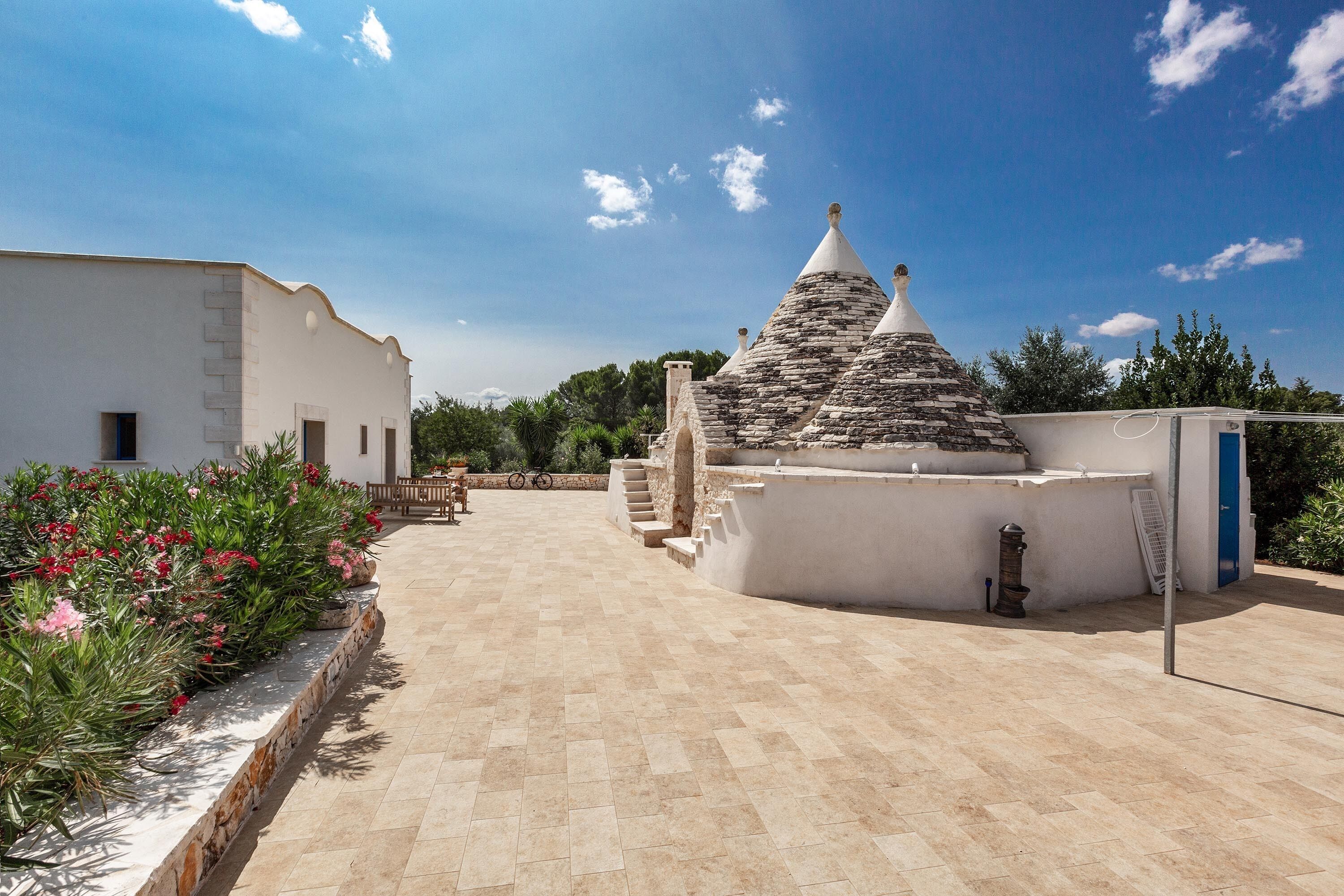 Un patio con un edificio blanco, una construcción de piedra con techo cónico y un sendero bordeado de plantas.