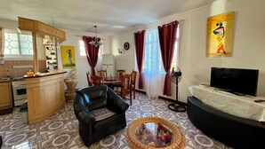 A living room with a black leather sofa, a wicker chair, a dining table with chairs, a TV on a stand, and a kitchen area with a stove and cabinets. - Villa Poisson dans L'eau (Trou-aux-Biches)