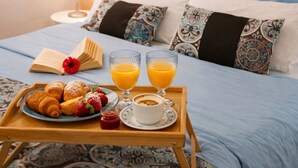 Uma cama bem arrumada com almofadas estampadas, uma bandeja com croissants, morangos e suco de laranja, e um abajur na mesinha de cabeceira.