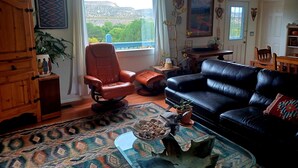 A living room with a black leather sofa, a brown leather chair, a wooden cabinet, a patterned rug, and a view of a mountainous landscape through a large window. - Cliff View Hacienda - Wild & Serene (Camp Verde)