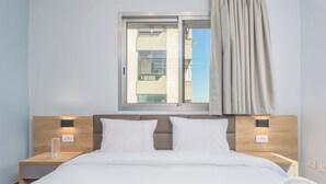 A hotel room with a bed, two bedside tables, a window with curtains, and a view of a building outside. - Grey - Elegant Apartments in Awkar (Mazraet El Hadira)