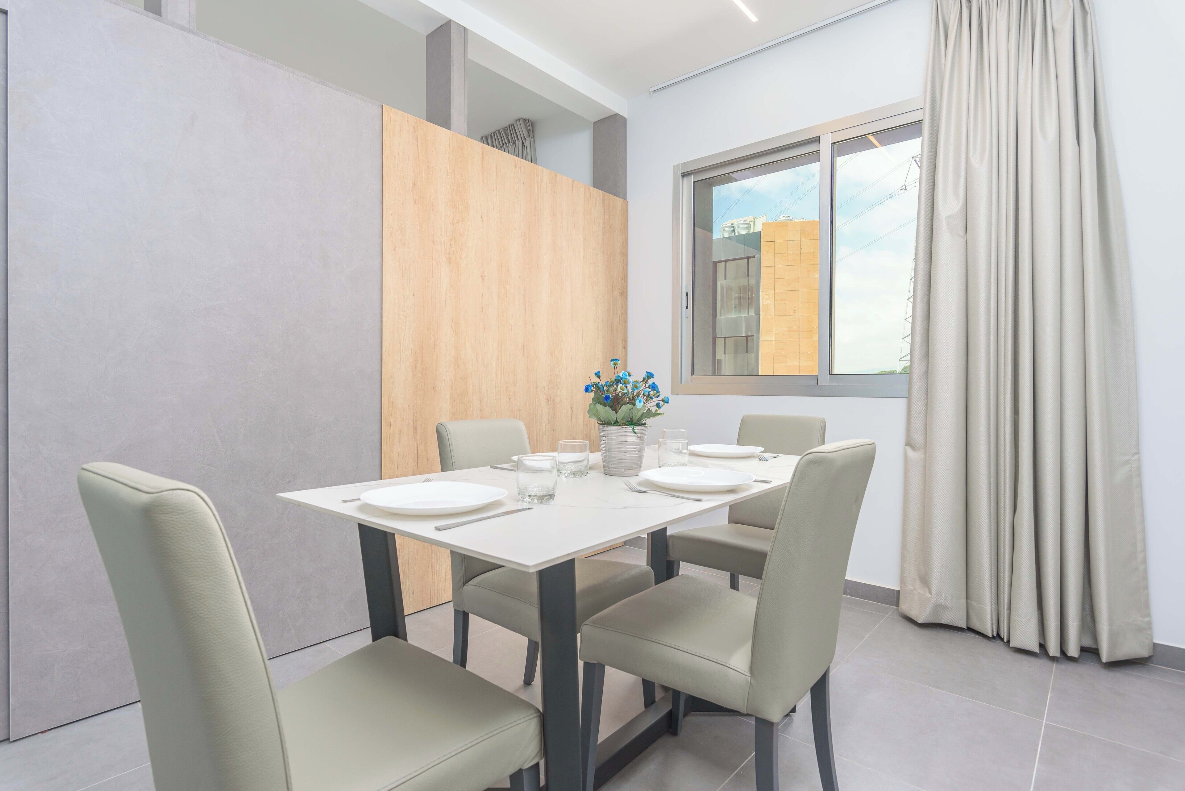 Een eetgedeelte met een witte tafel voor vier personen, beige stoelen en een vaas met bloemen.