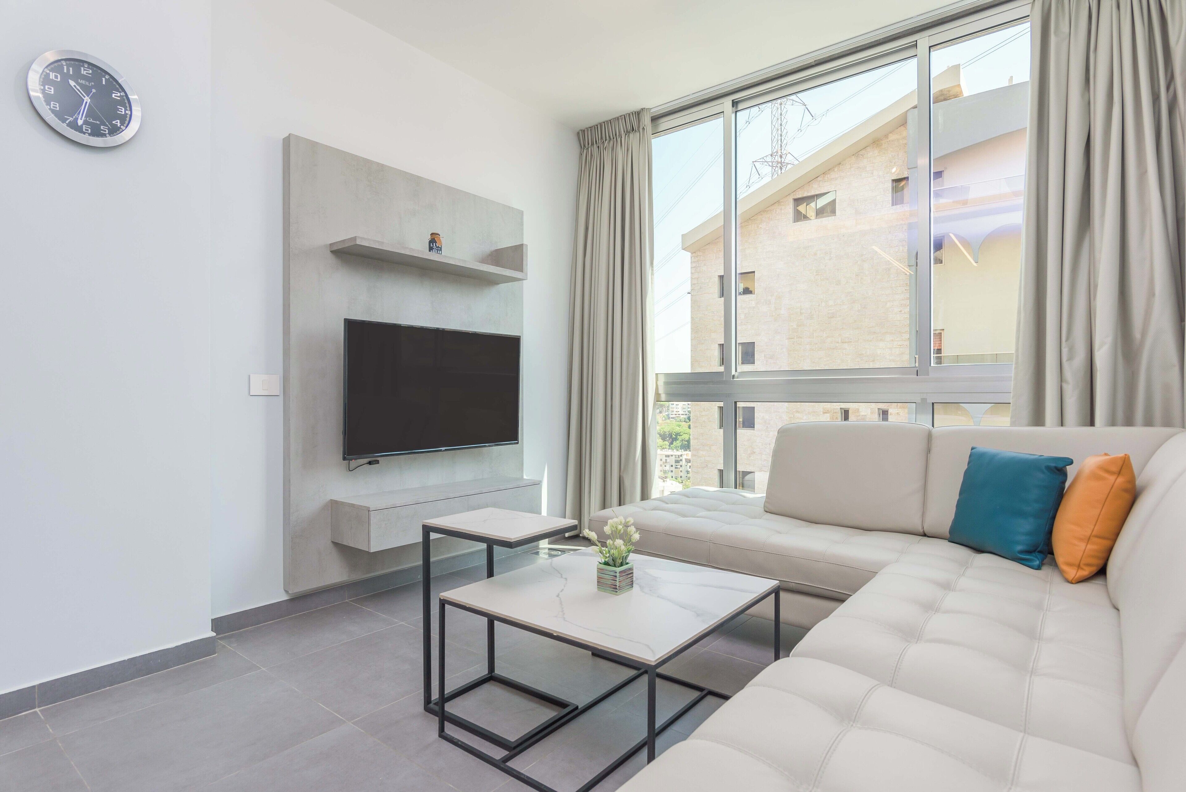A modern living room with a flat-screen TV, a sofa, a coffee table, and a large window with curtains.