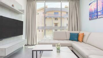 A modern living room with a flat-screen TV, a sofa with cushions, a coffee table, and a large window with curtains.