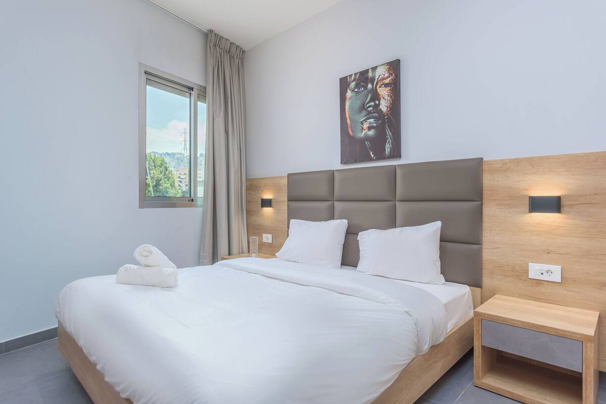 A modern hotel room with a large bed, a headboard with built-in lighting, a bedside table with a lamp, and a window with curtains.