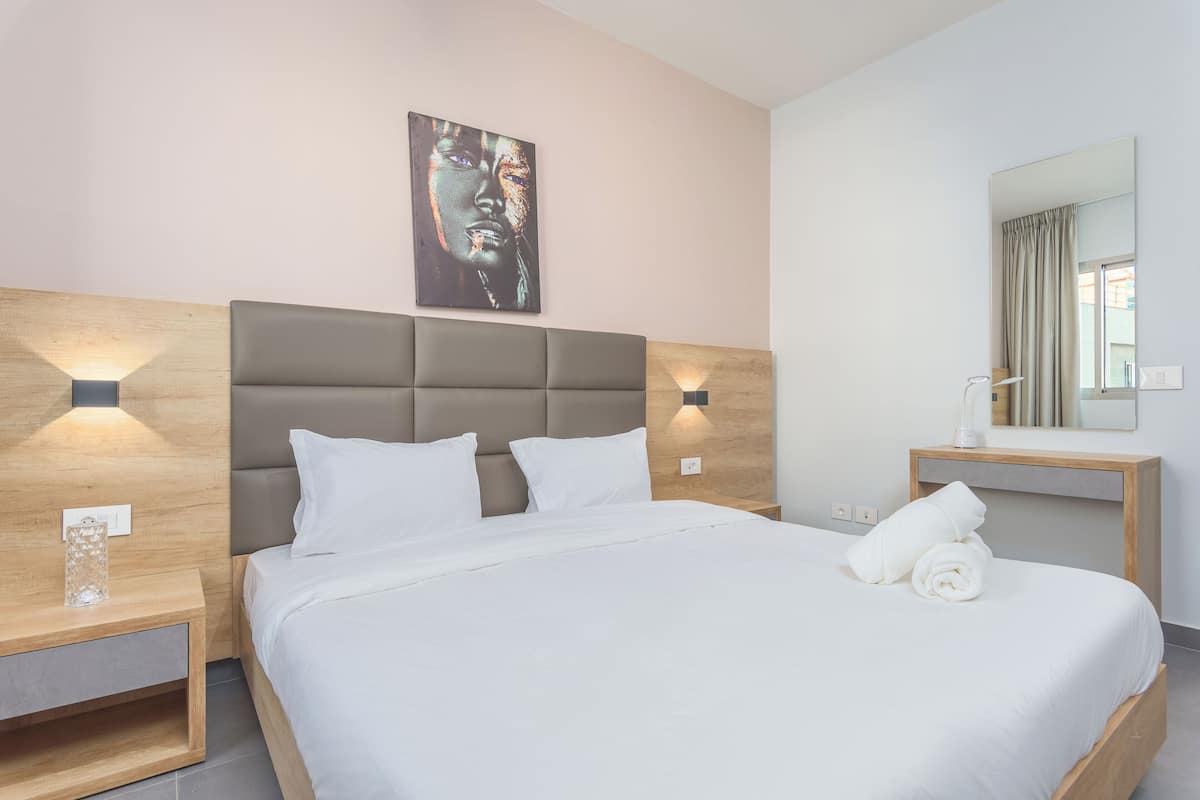 A modern hotel room with a large bed, a headboard with integrated lighting, a bedside table with a lamp, and a large mirror above the bed.