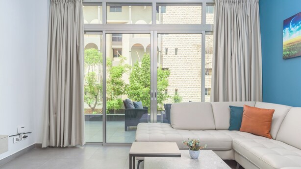 A modern living room with a beige sofa, a coffee table, and a sliding glass door leading to an outdoor area with a patio and greenery. - Grey - Elegant Apartments in Awkar (Mazraet El Hadira)
