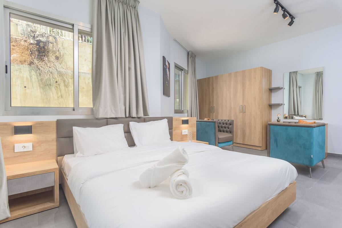 A modern hotel room with a large bed, a wooden wardrobe, a vanity with a mirror, and a view of a garden through the window.