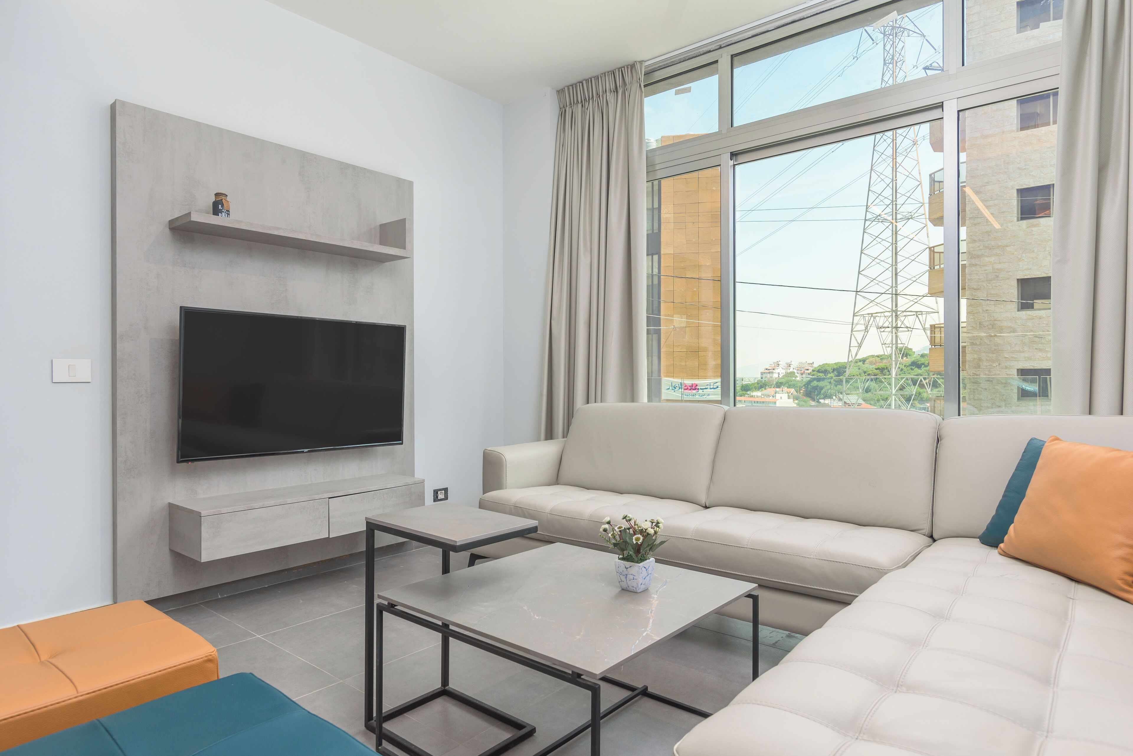 Een moderne woonkamer met een grote flatscreen televisie aan een wand, een beige bank, een salontafel en uitzicht op een gebouw door grote ramen.