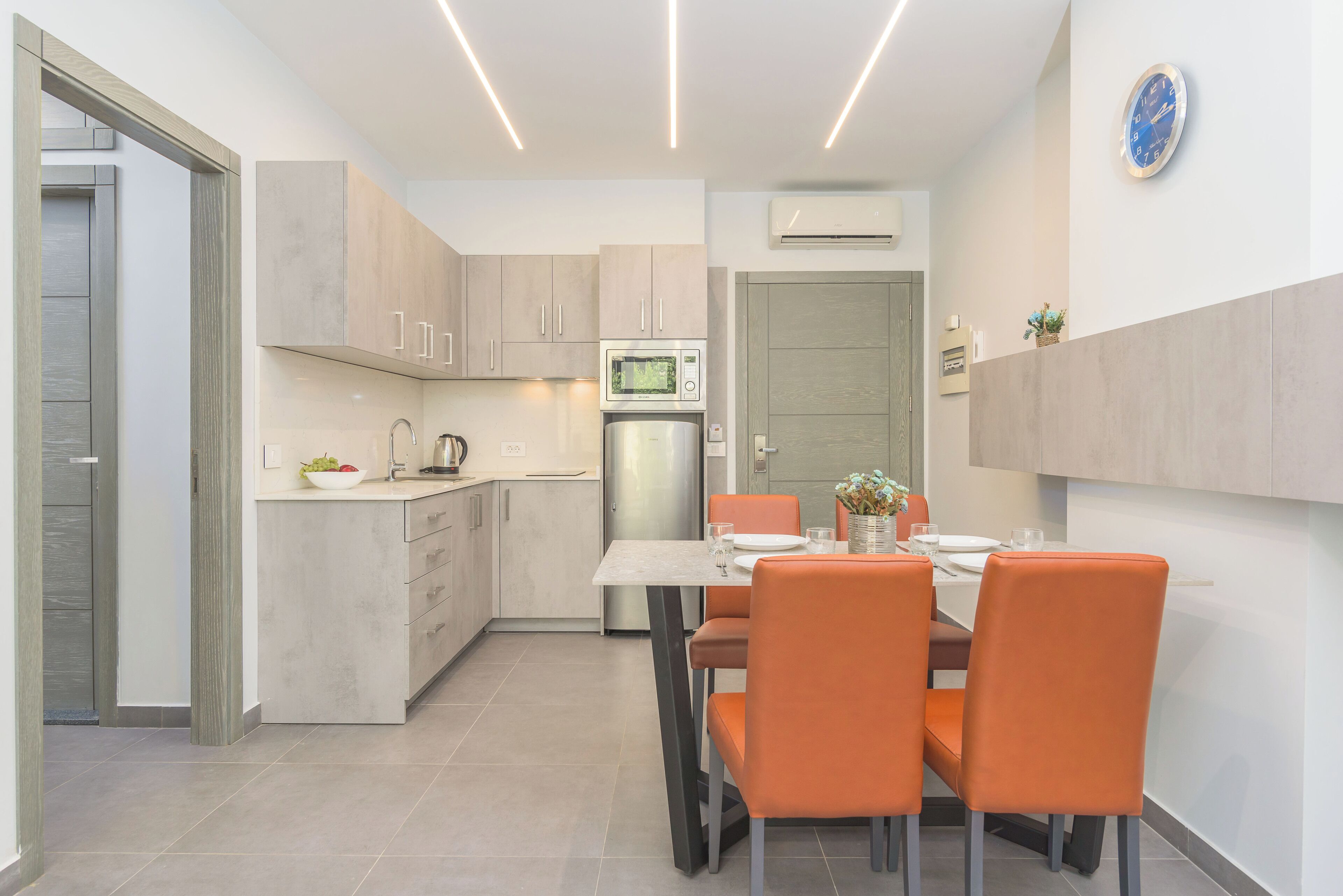 Una cocina moderna con zona de comedor, que incluye un microondas, refrigerador y una mesa de comedor con sillas naranjas.