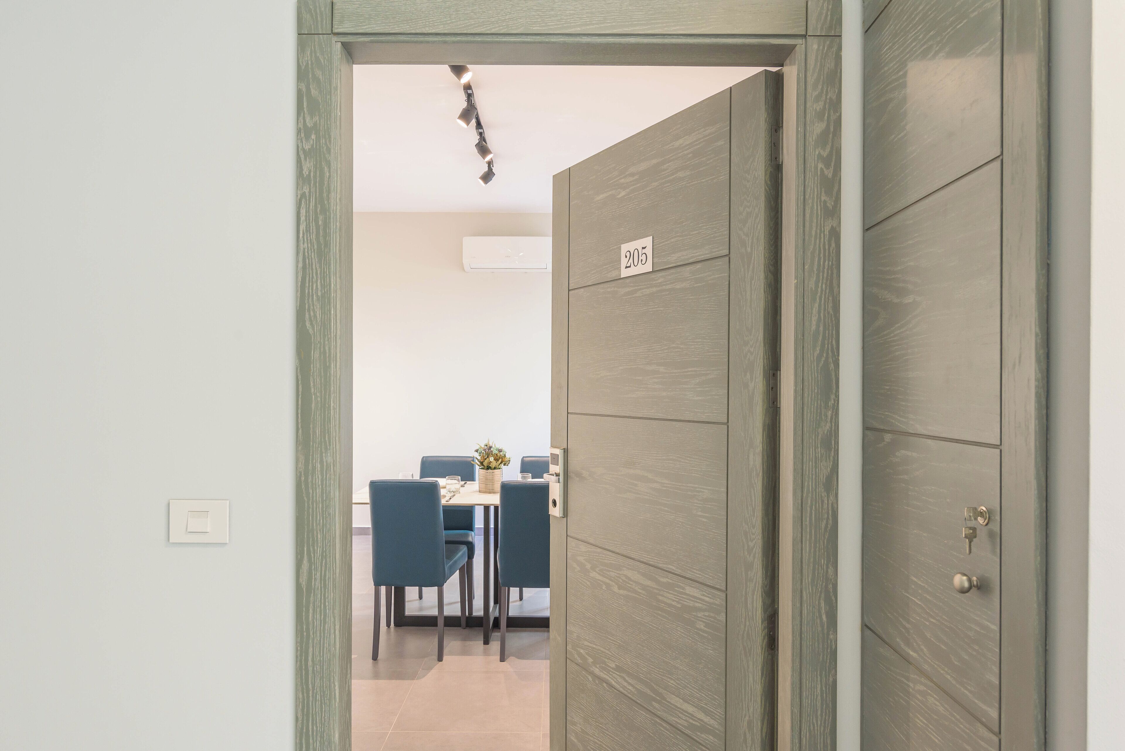 A room with a wooden door labeled "205," a dining area with blue chairs and a table, and a light switch on the wall.