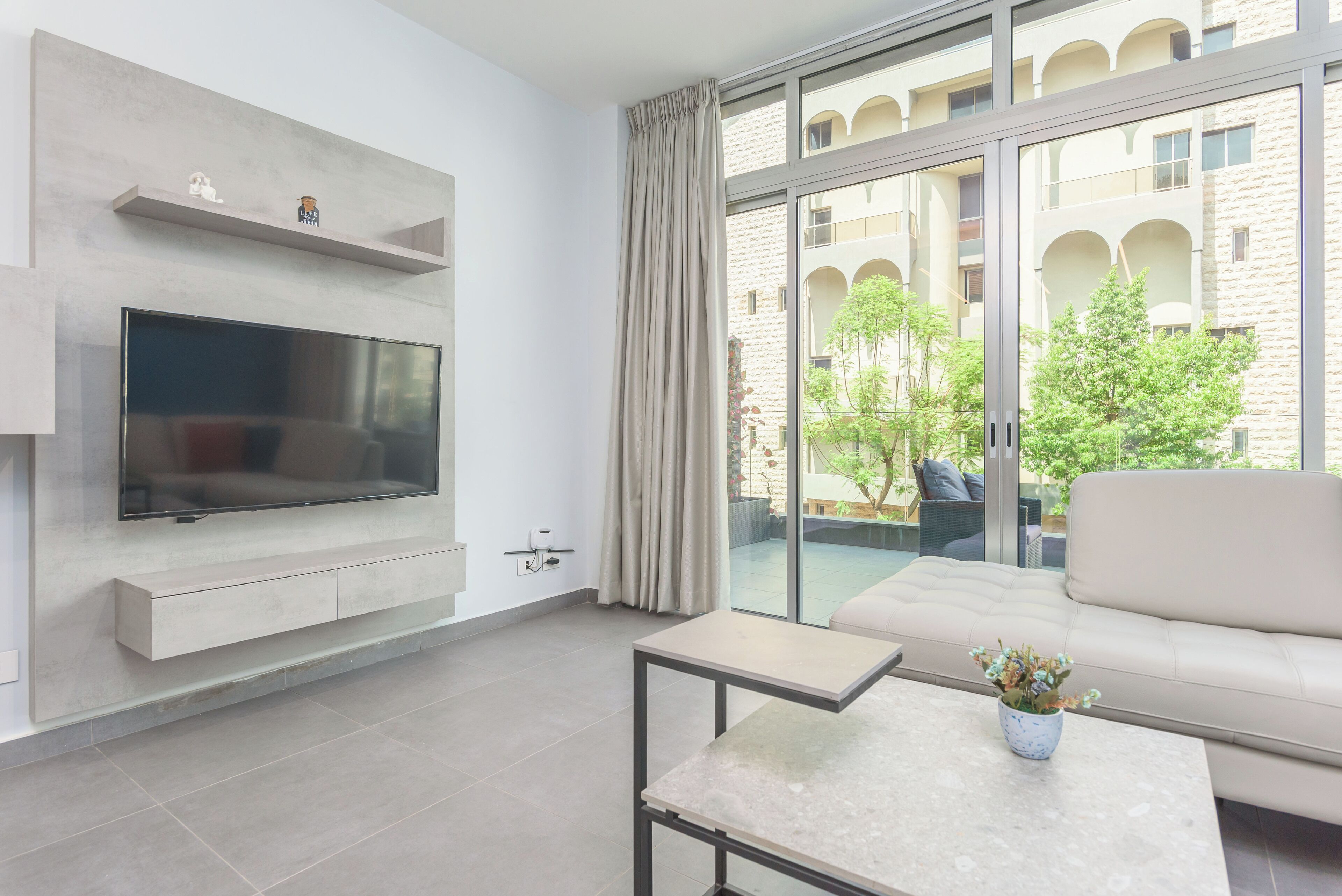 A modern living room with a flat-screen TV, a sofa, a coffee table, and a balcony with a view of a building and greenery.